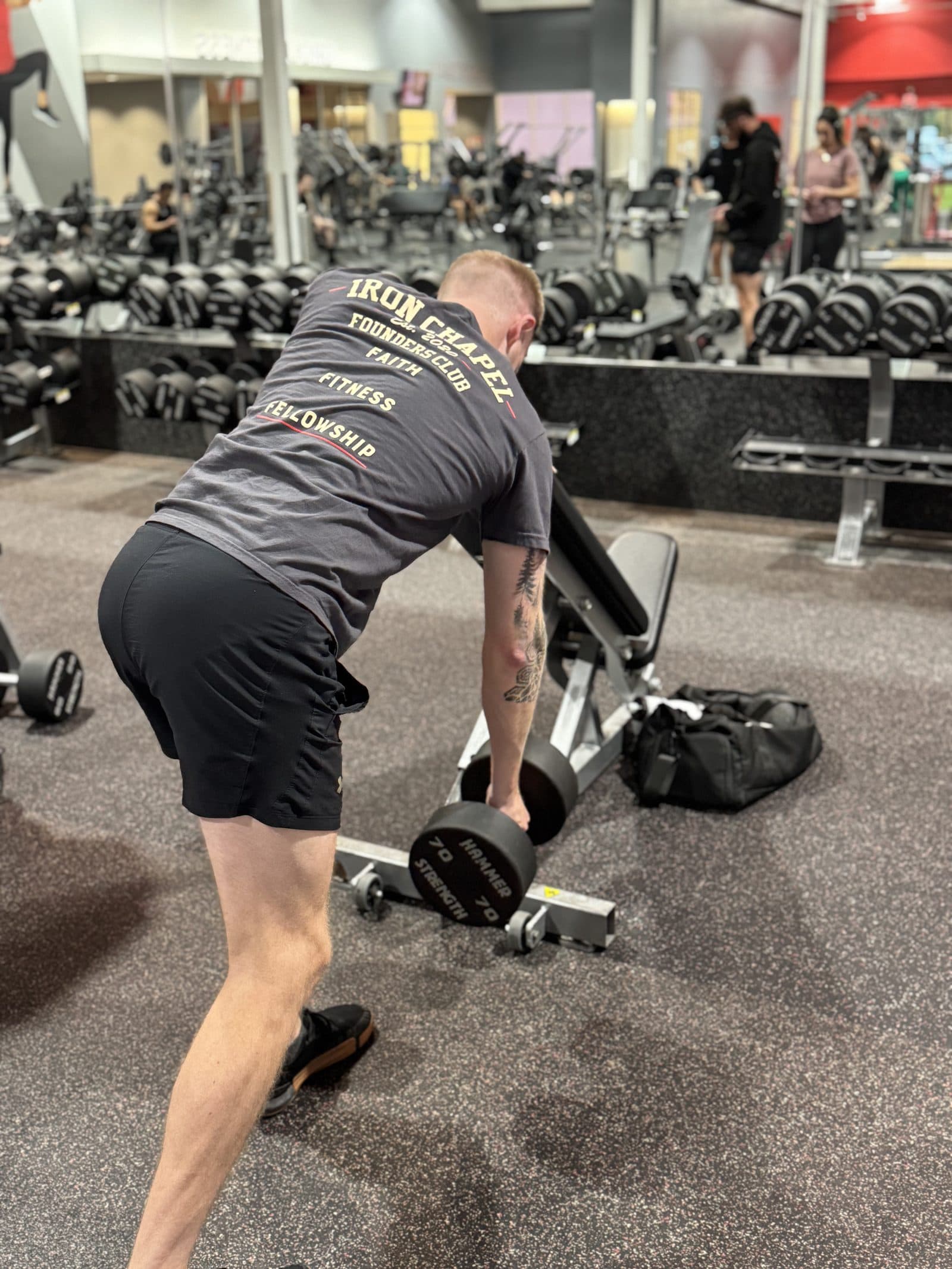 Founders Club tee worn from the back, rowing a dumbbell in the gym